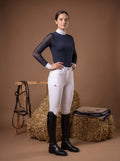 Woman in equestrian attire standing in front of a brown background with hay and a saddle.