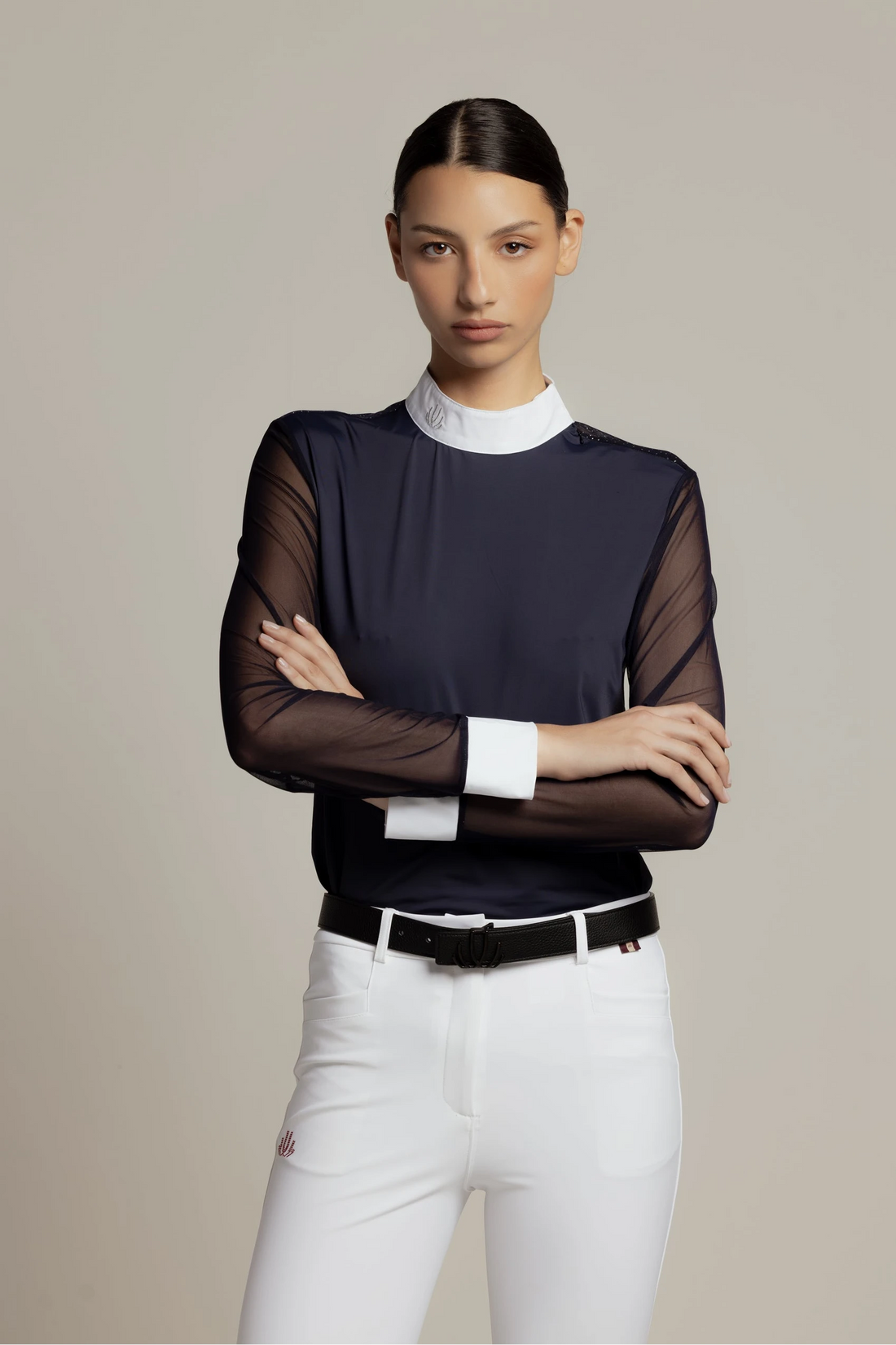 A model wearing a dark navy long-sleeve top with sheer mesh sleeves, a white mock neck collar, and white cuffs, paired with white pants and a black belt, against a solid beige background.
