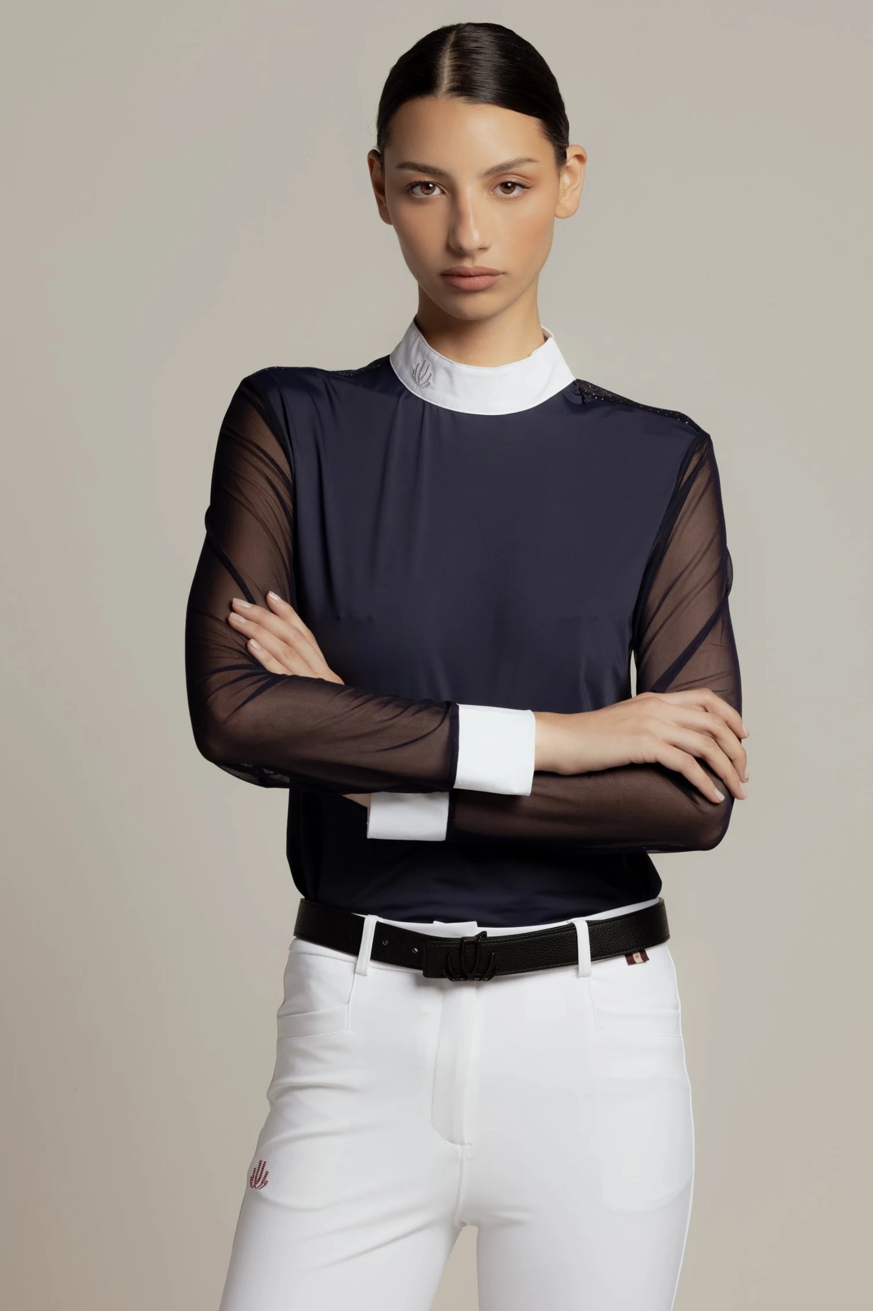 A model wearing a dark navy long-sleeve top with sheer mesh sleeves, a white mock neck collar, and white cuffs, paired with white pants and a black belt, against a solid beige background.