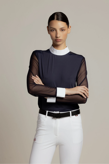 A model wearing a dark navy long-sleeve top with sheer mesh sleeves, a white mock neck collar, and white cuffs, paired with white pants and a black belt, against a solid beige background.