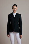 A woman modeling a black equestrian-style blazer with white piping on the lapels, worn over a white mock neck top and white riding breeches, against a solid beige studio background.