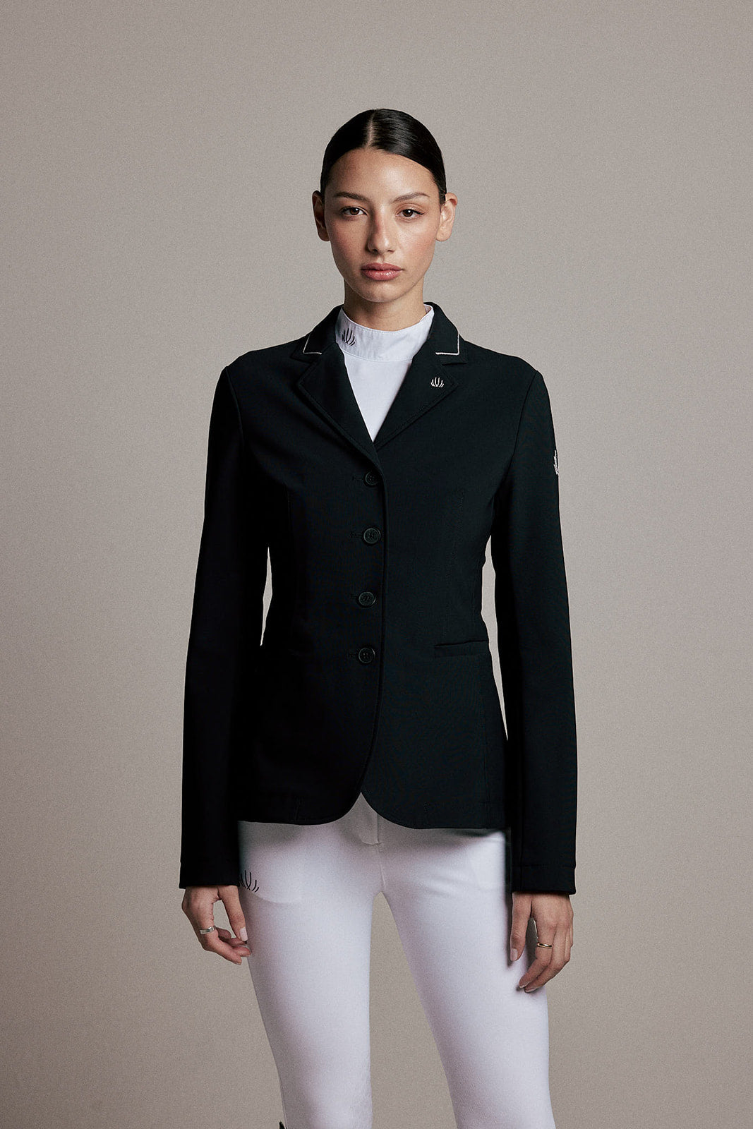 A woman modeling a black equestrian-style blazer with white piping on the lapels, worn over a white mock neck top and white riding breeches, against a solid beige studio background.