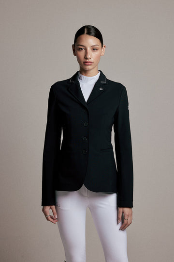 A woman modeling a black equestrian-style blazer with white piping on the lapels, worn over a white mock neck top and white riding breeches, against a solid beige studio background.