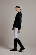 A model is shown in profile wearing a black equestrian-style blazer with white piping, white breeches, and black riding boots against a solid beige studio background.
