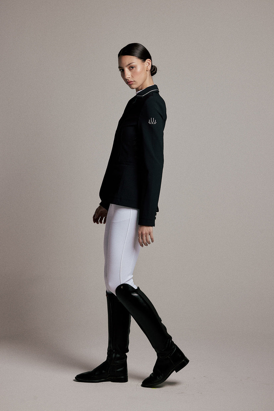A model is shown in profile wearing a black equestrian-style blazer with white piping, white breeches, and black riding boots against a solid beige studio background.