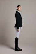 A woman in side profile wearing a black equestrian-style blazer, white breeches, and black riding boots against a plain taupe studio background.