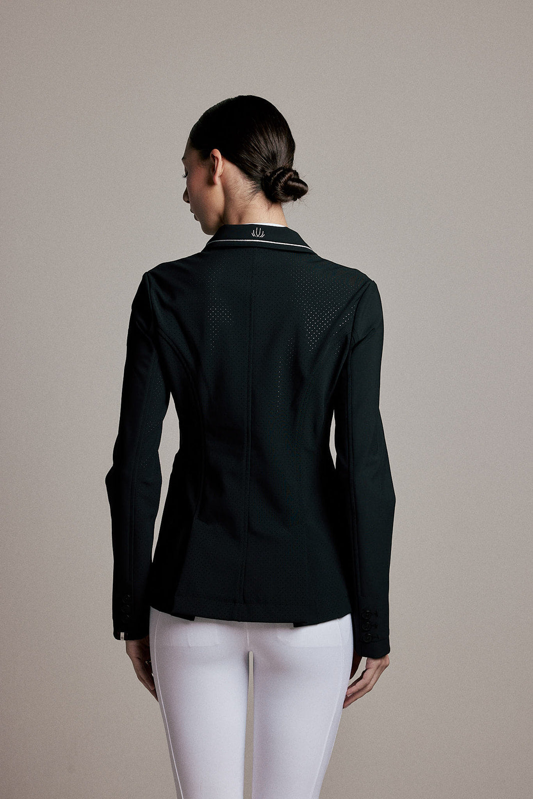 A woman shown from the back wearing a dark green fitted jacket with a subtle textured pattern on the back and white pants, set against a plain beige studio background.
