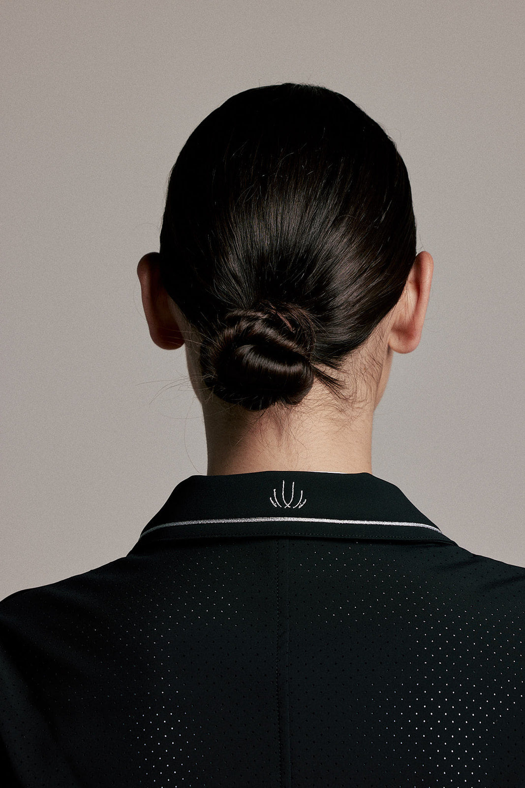 Back view of a person with dark hair in a low bun, wearing a black collared shirt with a small white embroidered logo on the collar, against a plain light gray background.