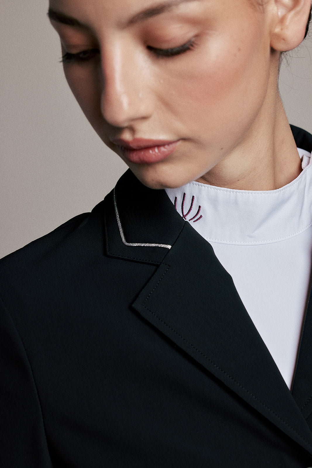 Close-up of a model wearing a black structured blazer with white contrast piping over a white mock-neck top, against a neutral beige background.