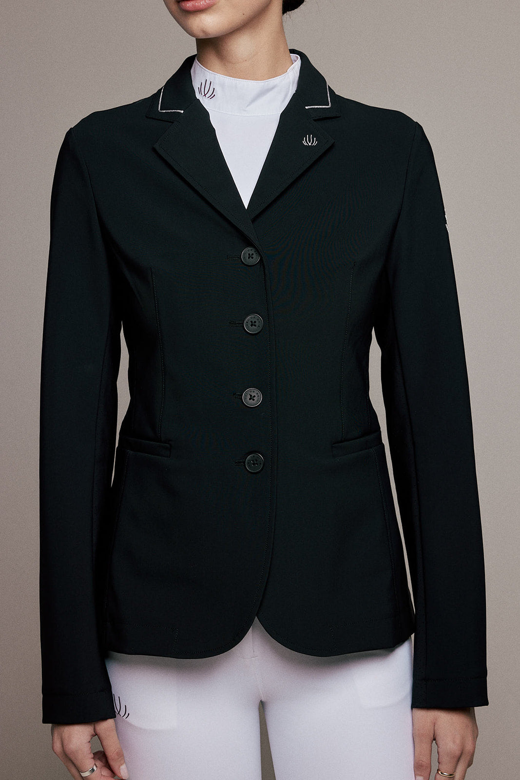 A woman wearing a tailored black equestrian-style blazer with white piping on the collar, a white mock neck top, and white riding pants, shown against a plain taupe studio background.