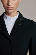 Close-up of a model wearing a black structured blazer with white piping on the collar and lapels, layered over a white mock-neck top. A small embroidered logo is visible on the left lapel.