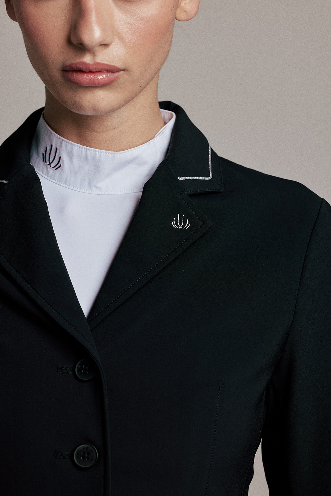 Close-up of a model wearing a black structured blazer with white piping on the collar and lapels, layered over a white mock-neck top. A small embroidered logo is visible on the left lapel.