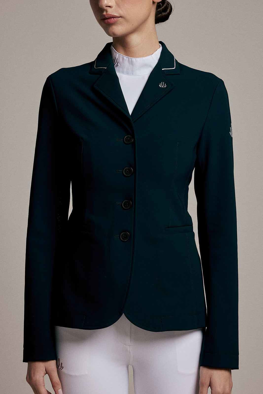 A woman wearing a dark teal equestrian blazer with white piping on the collar, a white ruffled mock neck, and white breeches, shown from the chin down against a neutral beige background.