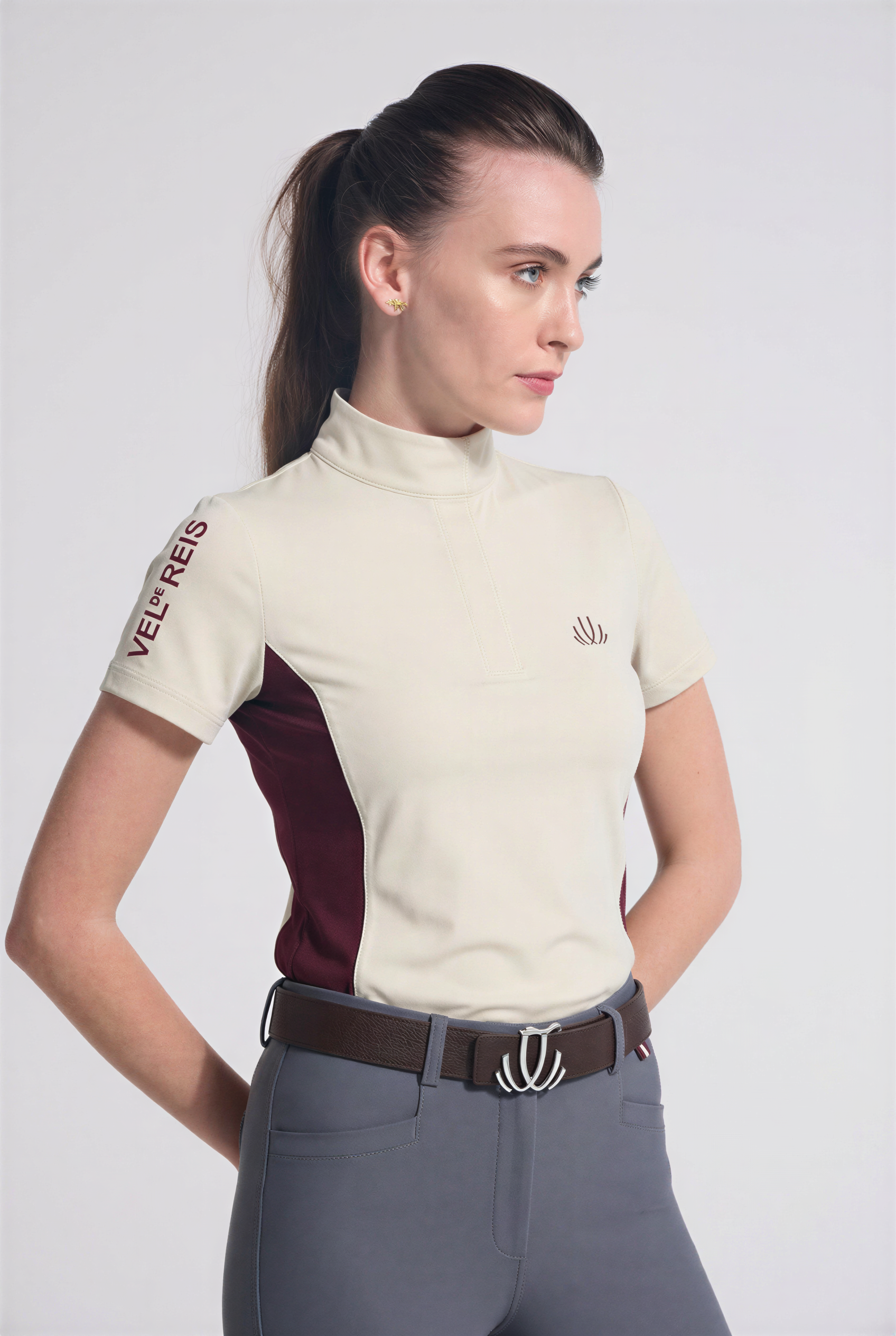 Woman wearing a beige equestrian training shirt with maroon accents and gray riding pants on a white background
